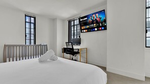 Una camera d'albergo con un letto, una scrivania con un computer, una televisione a parete e ampie finestre.