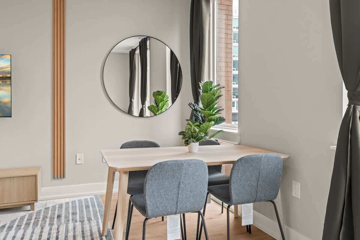 A dining area with a round mirror, a wooden table, four grey chairs, a potted plant, and a window with curtains.