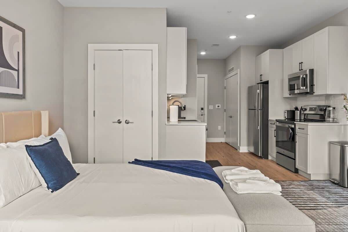 A modern hotel room with a large bed, a bedside table, a small seating area, and a kitchenette with a refrigerator, microwave, and oven.