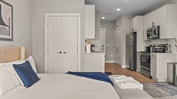 A modern hotel room with a large bed, a bedside table, a small seating area, and a kitchenette with a refrigerator, microwave, and oven.