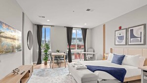 A modern hotel room with a bed, a desk, a chair, and a view of the city. - Luxury Studio | 1 Bed | Northern Liberties (Philadelphia)