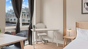 Ein Hotelzimmer mit Bett, Schreibtisch, Stuhl, einem kleinen Tisch mit Lampe und einem Fenster mit Vorhängen.