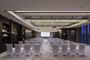 Uma sala de conferências com fileiras de cadeiras brancas, uma tela grande e um lustre moderno.
