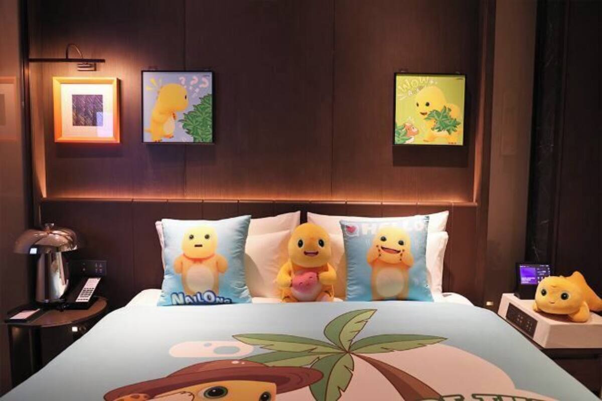 Habitación de hotel con cama, almohadones con personajes de dibujos animados, mesita de noche con lámpara y cuadros en la pared.