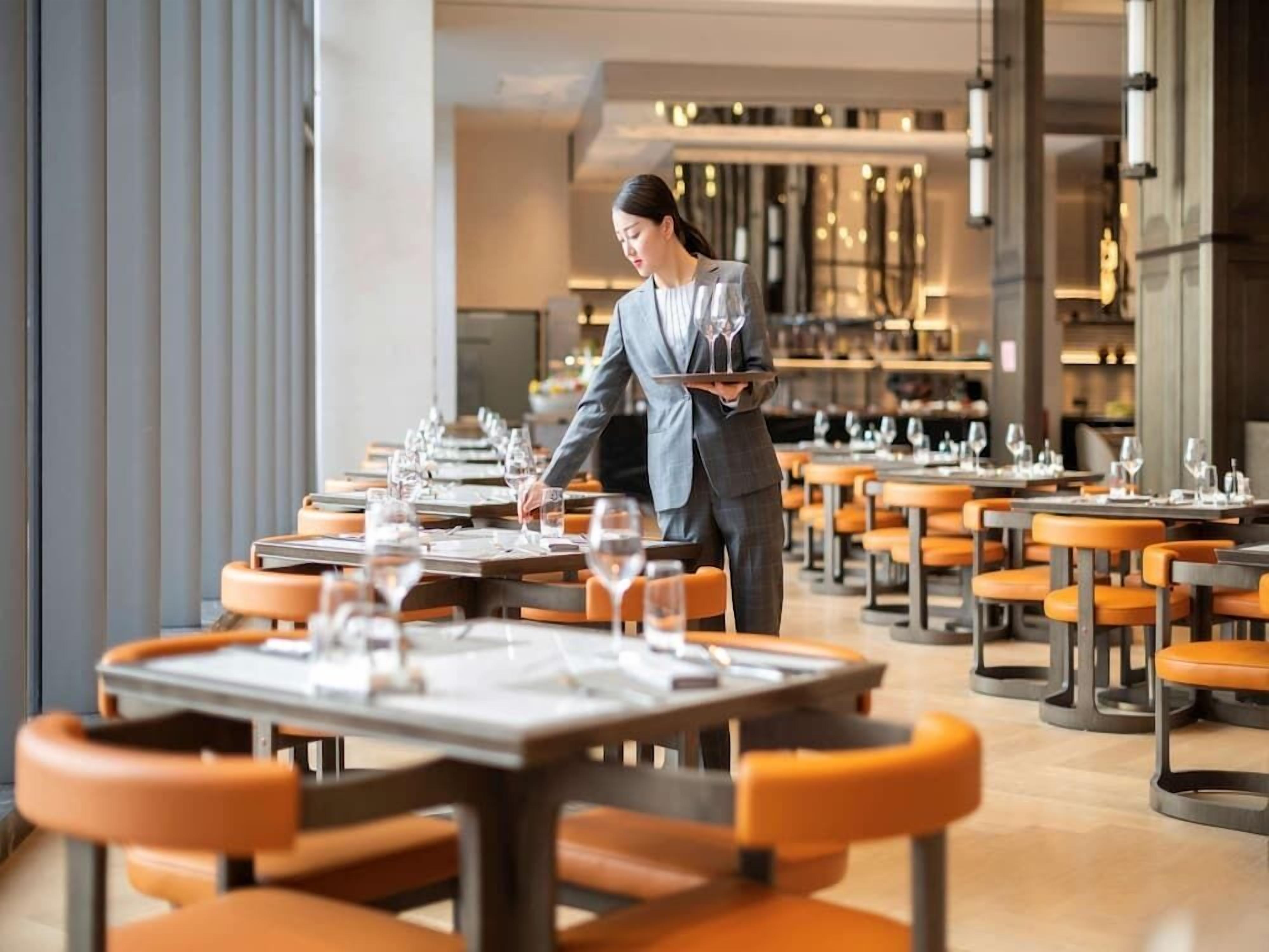 A restaurant with multiple tables and chairs, a waiter carrying a tray with glasses.