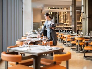 A restaurant with multiple tables and chairs, a waiter carrying a tray with glasses. - Novotel Xian Xixian (Xianyang)