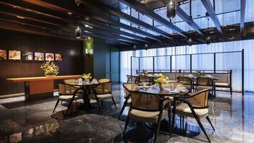 A modern dining area with high ceilings, exposed beams, and a mix of round and square tables with wicker chairs.