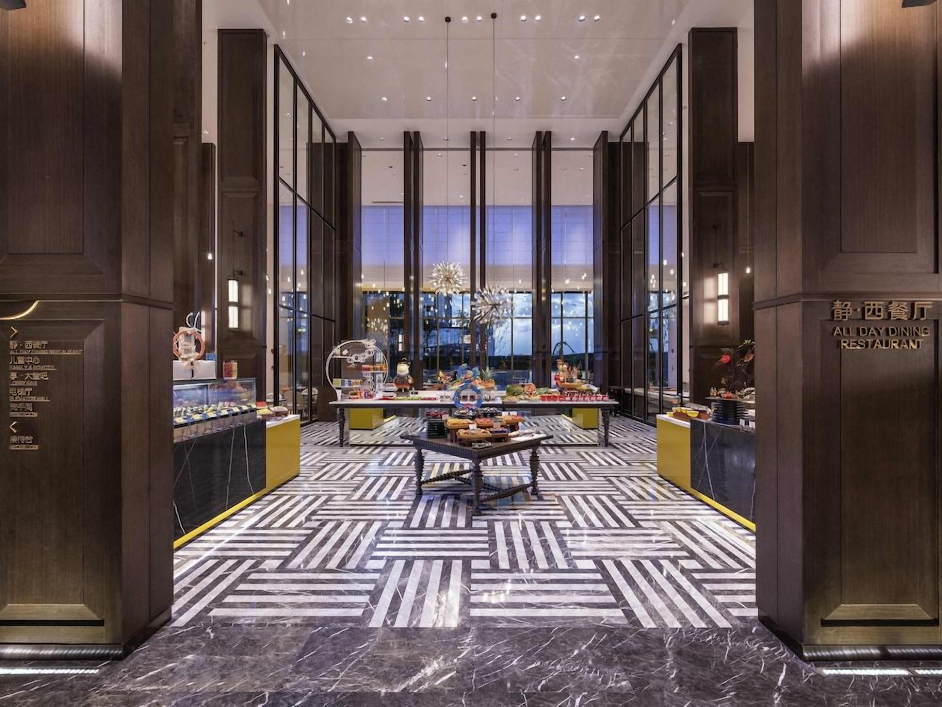 A modern hotel lobby with a central dining area, marble flooring, and a view of the cityscape through large windows.
