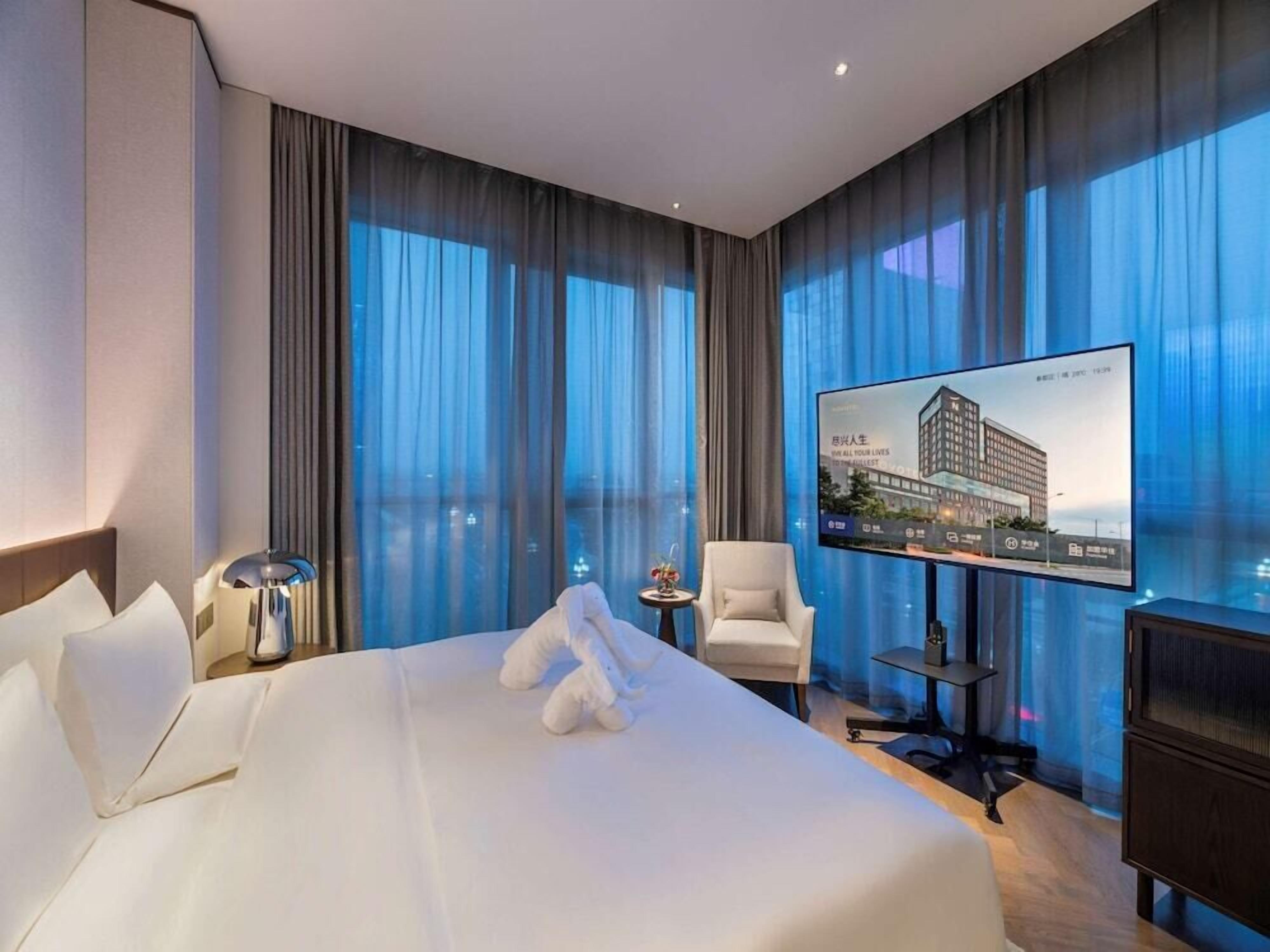 Ein Hotelzimmer mit einem großen Bett, einem flachbildfernseher an der Wand, einem kleinen Tisch mit einer Lampe, einem Sessel und Blick auf ein Gebäude durch das Fenster.