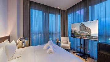 Ein Hotelzimmer mit einem großen Bett, einem flachbildfernseher an der Wand, einem kleinen Tisch mit einer Lampe, einem Sessel und Blick auf ein Gebäude durch das Fenster.