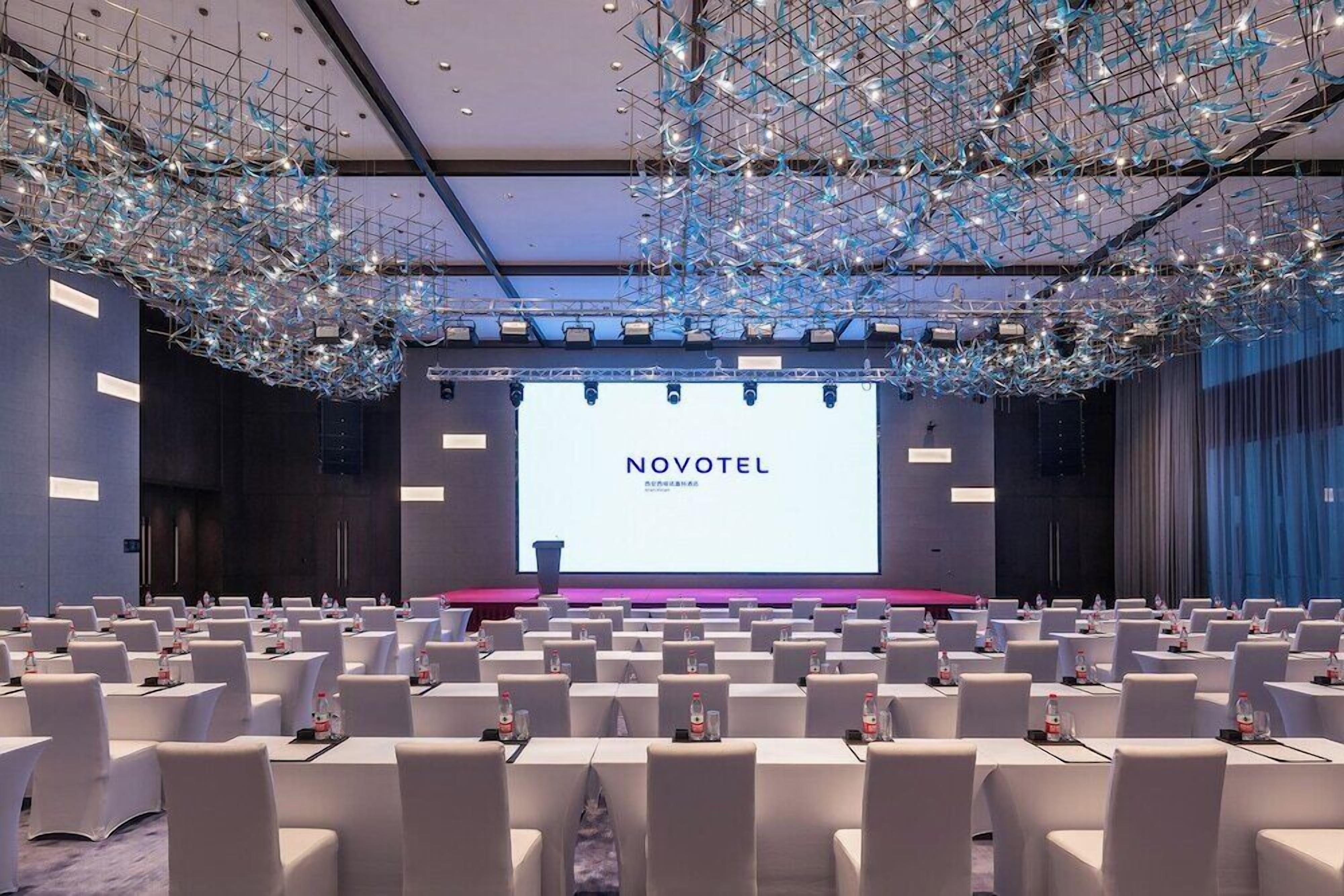 A conference room with rows of white tables and chairs, a large screen displaying 'NOVOTEL', and a modern ceiling design.