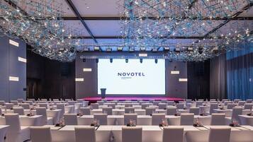 A conference room with rows of white tables and chairs, a large screen displaying 'NOVOTEL', and a modern ceiling design.