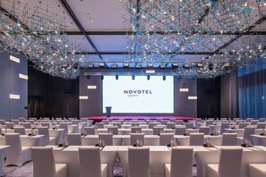 Une salle de conférence avec des rangées de tables et de chaises blanches, un grand écran affichant le mot « NOVOTEL », et un design moderne au niveau du plafond.