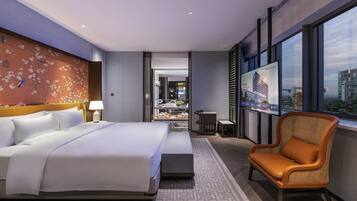 Habitación de hotel con una cama grande, zona de estar con una silla, televisor montado en la pared y vistas a la ciudad a través de amplios ventanales.