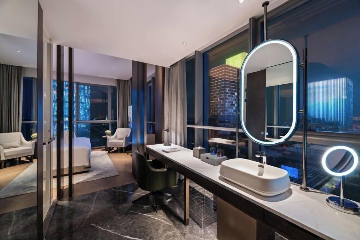 Un baño de hotel moderno con un espejo grande, un lavamanos y vistas a la ciudad.