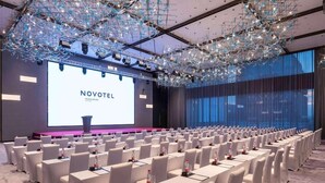 Une salle de conférence avec des rangées de chaises et de tables blanches, un grand écran affichant le mot « NOVOTEL », et un lustre moderne.