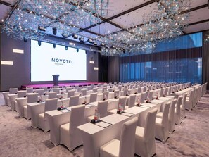 A conference room with rows of white chairs and tables, a large screen displaying 'NOVOTEL,' and a modern chandelier. - Novotel Xian Xixian (Xianyang)
