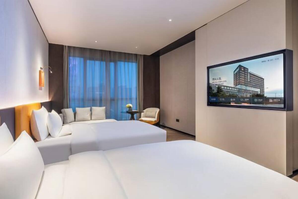 Ein Hotelzimmer mit zwei Betten, einem großen Fenster mit Vorhängen, einem kleinen Tisch mit einem Stuhl und einem an der Wand montierten Fernseher, der ein Bild eines Gebäudes zeigt.