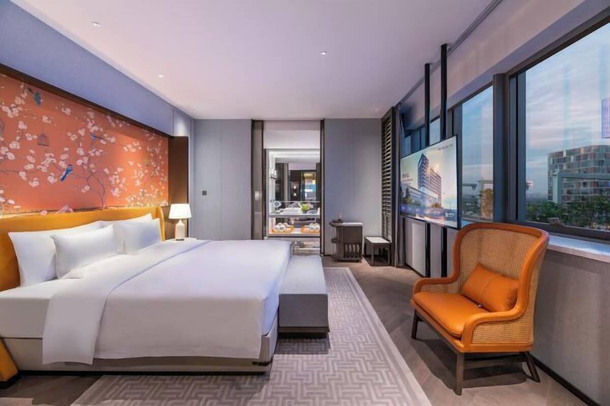 Ein Hotelzimmer mit Bett, Sessel, Wand mit Kunstwerk, Fenster und Blick auf die Stadt.