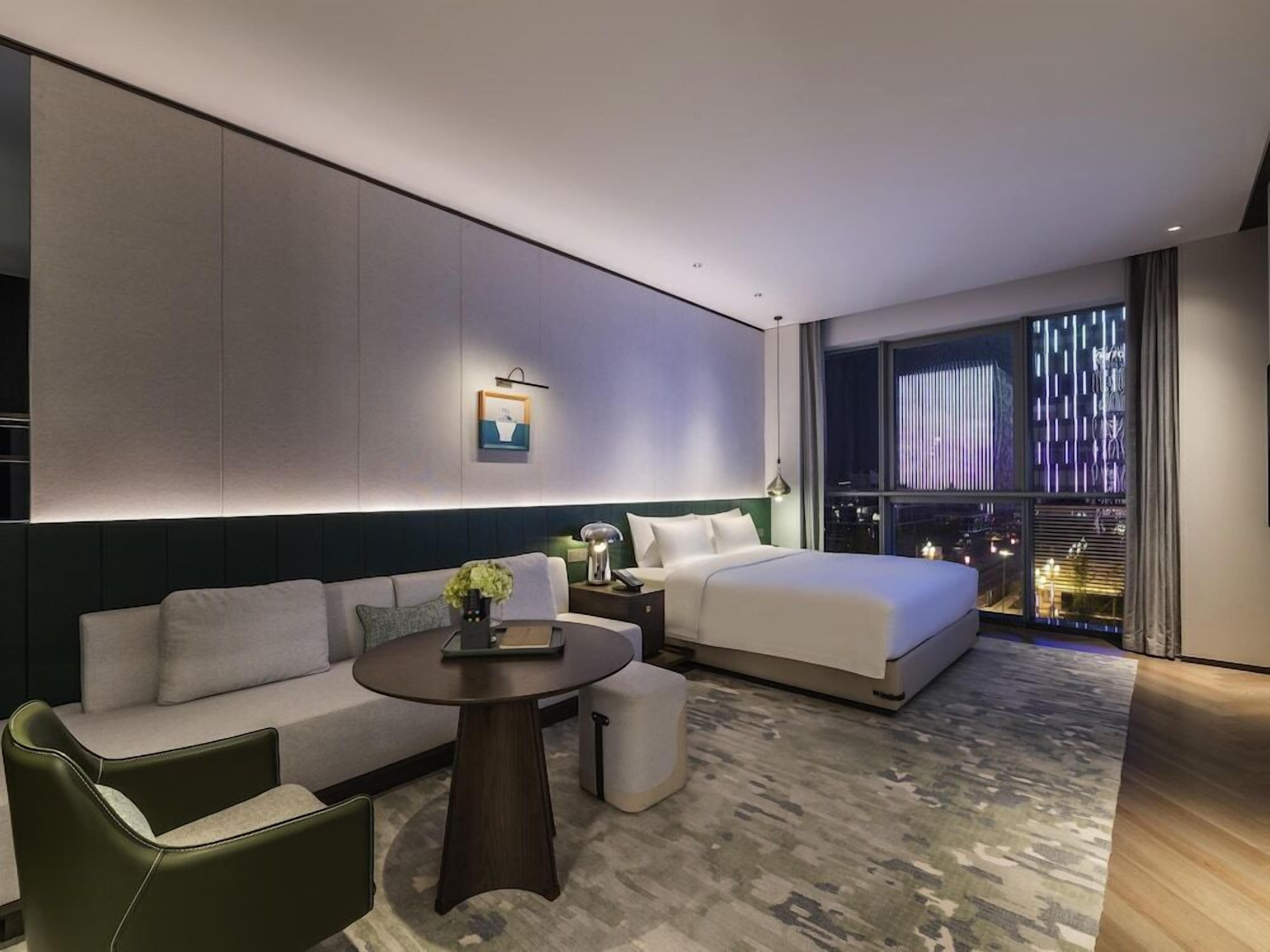 A modern hotel room with a bed, sofa, and a small table with a lamp.
