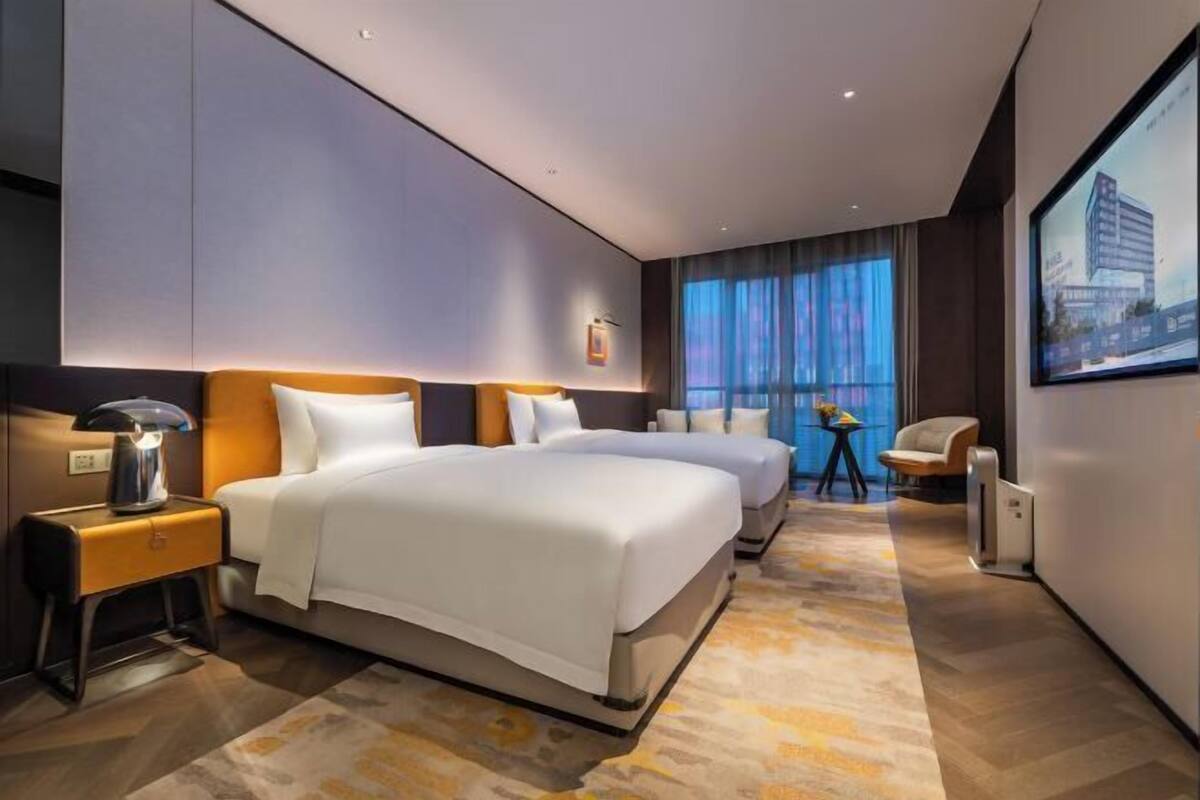 Ein Hotelzimmer mit zwei Betten, einem Nachttisch, einer Lampe, einem Sessel, einem Tisch und einem großen Fenster mit Vorhängen.