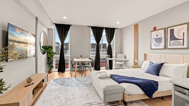 A modern hotel room with a large bed, a desk, a chair, and a view of the city. - Luxury Studio | 1 Bed | Northern Liberties (Philadelphia)