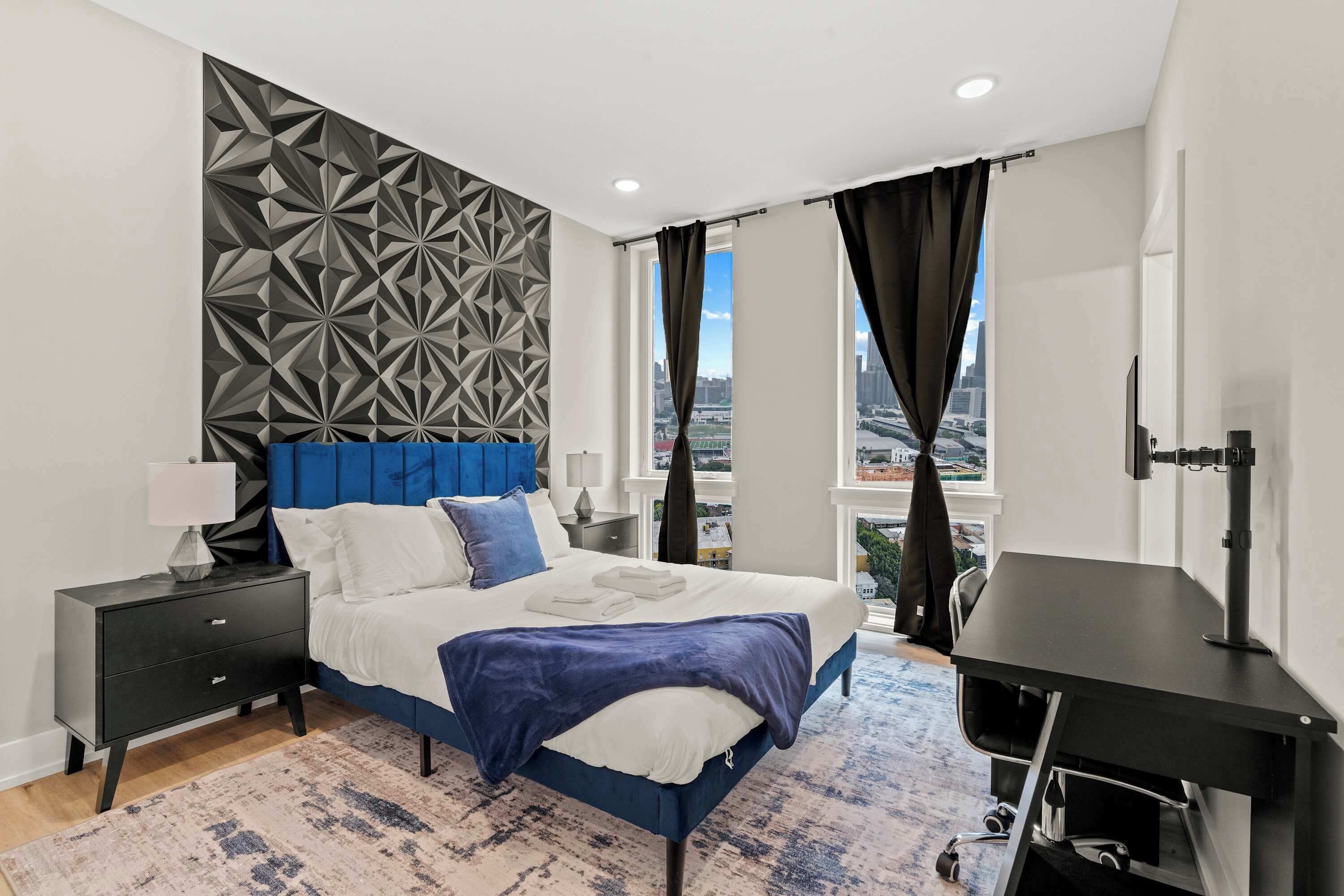Une chambre moderne avec une tête de lit à motifs géométriques, un lit avec une literie blanche et bleue, une table de chevet avec une lampe, un bureau avec un ordinateur et une vue sur le paysage urbain à travers de grandes fenêtres.