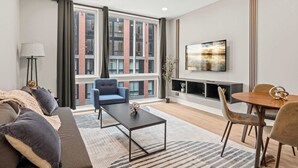 A modern living room with a sofa, a coffee table, a blue armchair, a dining table with chairs, a fireplace, and a large window with curtains.
