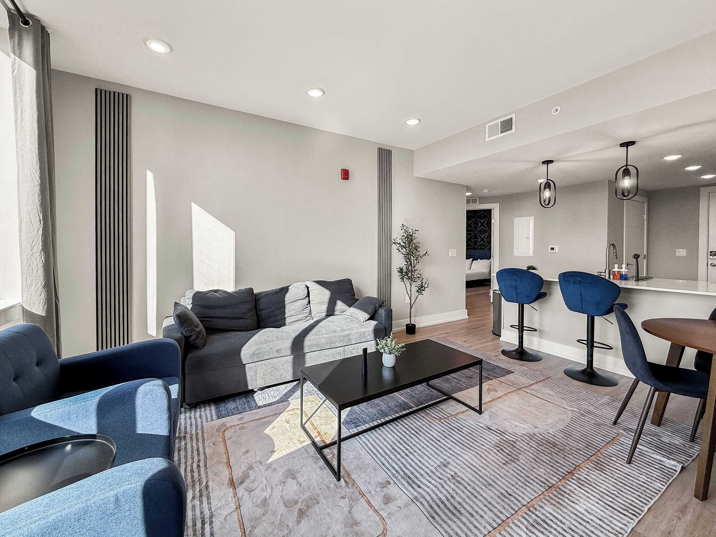 A modern living room with a sofa, coffee table, and blue chairs. There is a dining area with a table and chairs, and a kitchen in the background.
