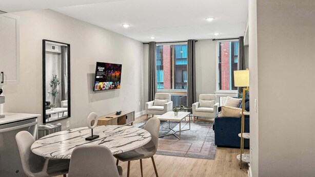 A modern hotel room with a dining area, a TV mounted on the wall, and large windows with curtains. - Luxury 1BD | 2 Beds | Northern Liberties (Philadelphia)