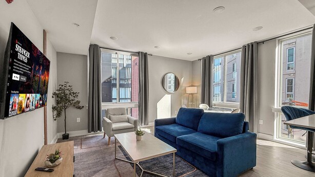 A modern living room with a blue sofa, a white armchair, and a coffee table. There is a TV mounted on the wall displaying a movie series. - Luxury 1BD | 2 Beds | Northern Liberties (Philadelphia)