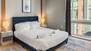A hotel room with a large bed, two bedside tables, a nightstand, a framed artwork on the wall, and a view of the city through the window. - Luxury 1BD | 2 Beds | Northern Liberties (Philadelphia)