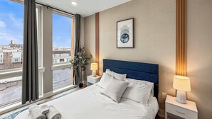 A hotel room with a large bed, two bedside tables, a nightstand, a window with curtains, a framed picture on the wall, and a city view. - Luxury 1BD | 2 Beds | Northern Liberties (Philadelphia)
