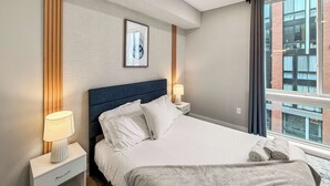 A modern hotel room with a large bed, two bedside tables, a framed picture on the wall, and a window with a city view.