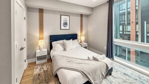 A modern hotel room with a large bed, two bedside tables, a wall-mounted artwork, and a large window with a city view.