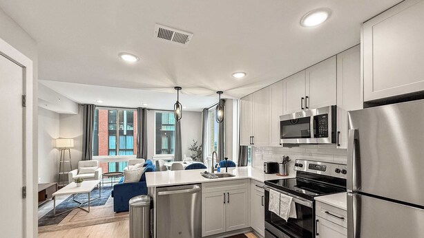 A modern kitchen with stainless steel appliances, a dining area with a blue sofa, and a view of a brick building through large windows.