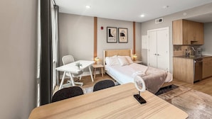 A modern hotel room with a bed, a desk, a chair, a small table, and a kitchenette.