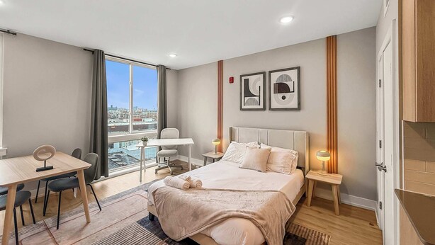 A modern hotel room with a large bed, a desk, and a chair, a view of the city through the window, and two framed artworks on the wall.