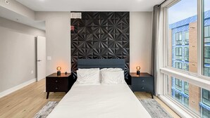A modern bedroom with a geometric-patterned headboard, a large bed, two bedside tables with lamps, and a view of a building through a large window. - Luxury 2BD | 4 Beds | Northern Liberties (Philadelphia)