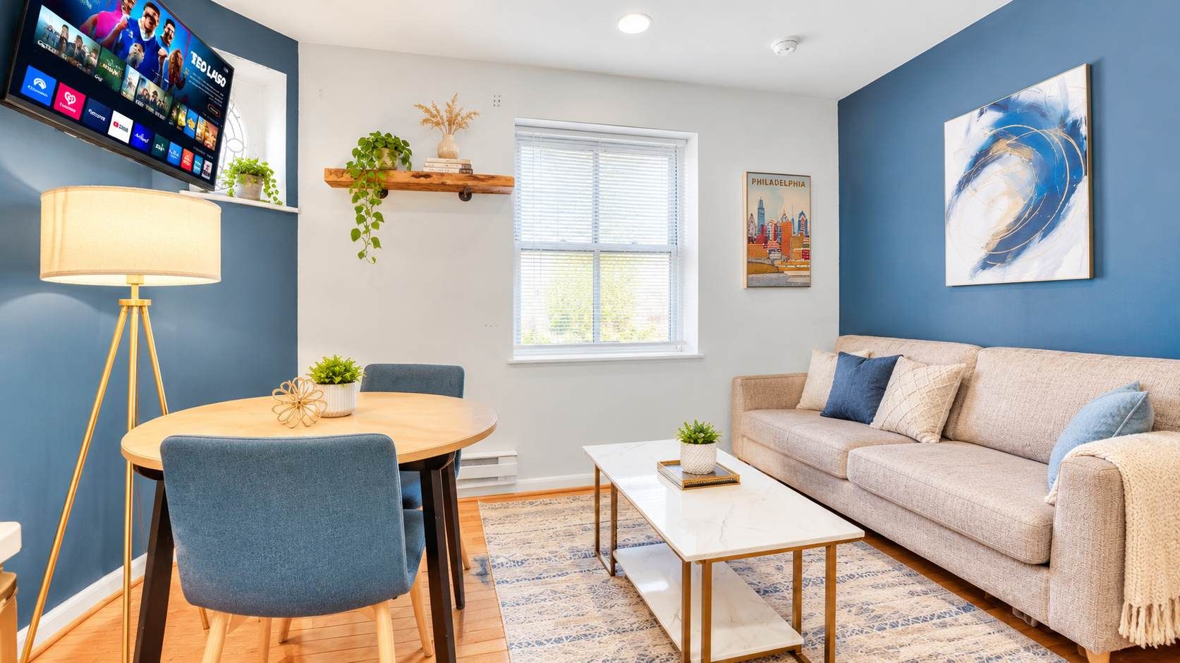 A living room with a sofa, coffee table, and chairs, a TV mounted on the wall, and a window with blinds.