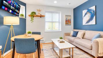 A living room with a sofa, coffee table, and chairs, a TV mounted on the wall, and a window with blinds.