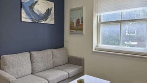 A living room with a grey sofa, a painting, a poster, and a window with a view of buildings. - Luxury 2BD | 5 Beds | Rittenhouse (Philadelphia)