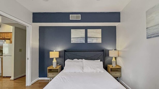 A bedroom with a large bed, two bedside tables with lamps, and a view of the kitchen through an open door. - Luxury 2BD | 5 Beds | Rittenhouse (Philadelphia)