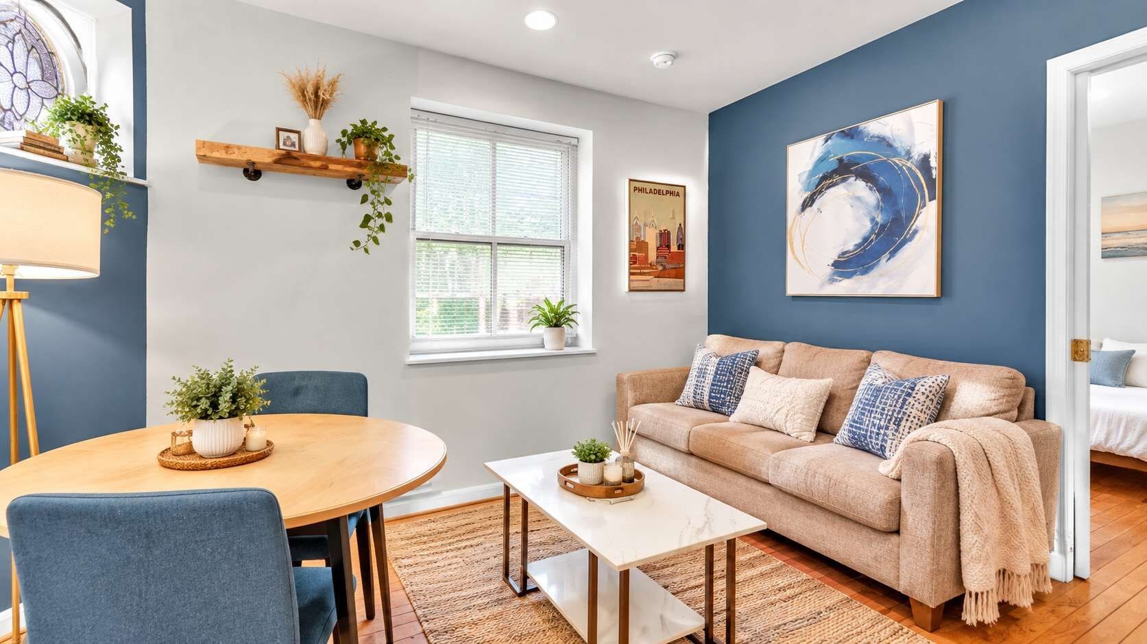 A living room with a round table, a sofa, and a coffee table. There are plants on shelves and a window with blinds.