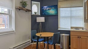 A compact living space with a round wooden table, two blue chairs, a small kitchen area, and a large window with blinds. - Luxury 2BD | 5 Beds | Rittenhouse (Philadelphia)