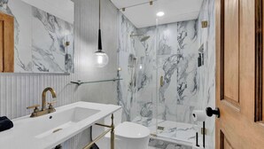 A modern bathroom with a marble backsplash, a glass shower enclosure, a white sink with gold fixtures, and a wooden door. - Luxury 1BD | 1 Bed | South St (Philadelphia)