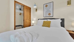 Ein ordentlich bezogenes Bett mit weißer Bettwäsche, eine Holztür, eine an der Wand montierte Lampe und ein gerahmtes abstraktes Kunstwerk.