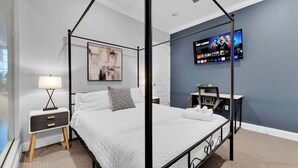 A four-poster bed with a white bedspread, a bedside table with a lamp, a TV mounted on the wall, and a desk with a chair.
