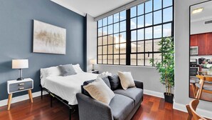 A modern bedroom with a bed, bedside tables, a grey sofa, a large window, and a view of the outside.