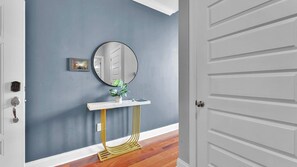 Un pasillo con pared azul, una puerta blanca, un espejo redondo, una pequeña consola blanca con base dorada y una planta en maceta.